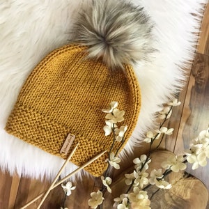 Pode incluir: Touca de malha amarelo mostarda com um grande pompom de pele sintética cinza. O chapéu tem uma bainha canelada e uma pequena etiqueta de madeira. Agulhas de tricô de madeira e flores brancas são dispostas perto do chapéu.