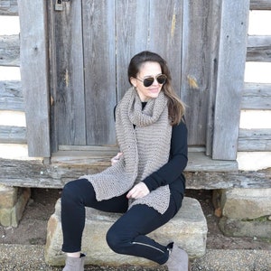 Op de afbeelding: Een lichtbruine gebreide sjaal over een zwarte top. De sjaal is rechthoekig en heeft een getextureerd uiterlijk. De persoon draagt een zwarte broek en grijze enkellaarsjes. De achtergrond is een verweerde houten deur.