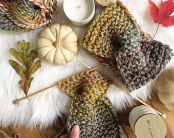 Könnte beinhalten: Ein handgestricktes Stirnband aus meliertem Garn in herbstlichen Farben wie Grün, Gelb und Braun. Das Stück ist im kraus rechts Muster gestrickt und hat ein zentrales gedrehtes Detail, präsentiert mit einem passenden Garnknäuel und Nadeln.