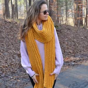 May include: A woman wearing a long, mustard yellow knitted scarf. The scarf is thick and chunky, and the woman is wearing it wrapped around her neck and down her chest. She is standing in a wooded area, and the background is blurred.