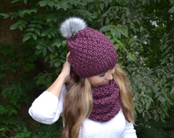 May include: A burgundy knitted hat with a faux fur pom pom and a matching burgundy infinity scarf.