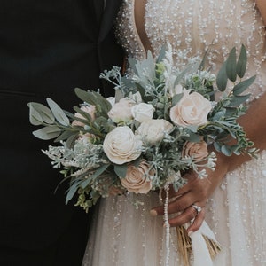 May include: A bride holds a bouquet of white and blush pink flowers with greenery. The bouquet is tied with a white ribbon and has a pearl strand hanging from it.