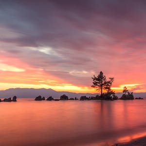 May include: A scenic landscape photograph of a lake at sunset. The sky is filled with vibrant colors of pink, orange, and purple. Silhouetted trees and rocks are in the water, with mountains in the distance.