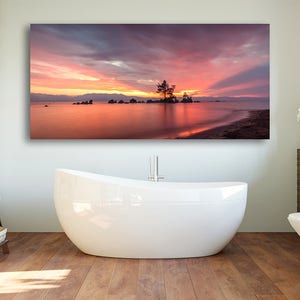 May include: A large rectangular canvas print of a sunset over a lake, with a small island and a tree silhouetted against the colorful sky. The water reflects the vibrant colors of the sunset. The print is displayed above a white bathtub in a bathroom.