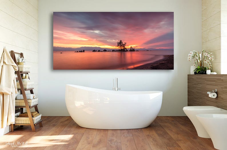 May include: A large rectangular canvas print of a sunset over a lake, with a small island and a tree silhouetted against the colorful sky. The water reflects the vibrant colors of the sunset. The print is displayed above a white bathtub in a bathroom.