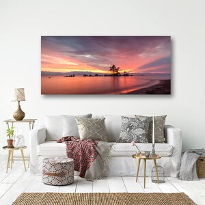 May include: A panoramic canvas print of a sunset over a lake. The sky is filled with shades of pink, orange, and purple. A small island with a tree is in the center of the lake. The image is displayed above a white sofa.