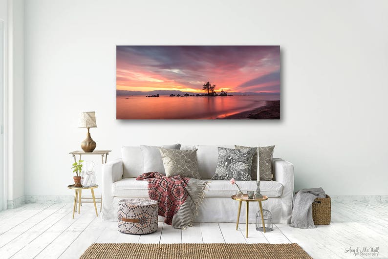 May include: A panoramic canvas print of a sunset over a lake. The sky is filled with shades of pink, orange, and purple. A small island with a tree is in the center of the lake. The image is displayed above a white sofa.