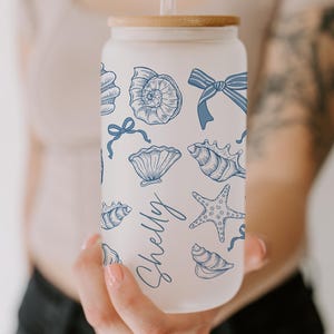 K&ouml;nnte beinhalten: Ein Glasbecher mit Bambusdeckel und Strohhalm, verziert mit blauen Muschel- und Schleifenillustrationen. Der Name "Shelly" ist in einer Schreibschrift geschrieben. Der Becher wird vor einer Person gehalten.