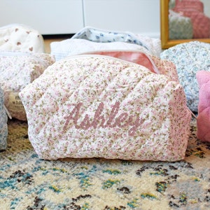 May include: A set of six quilted makeup bags with floral patterns. The bags are in various colors, including pink, blue, and white. One bag has the name "Ashley" embroidered on it in pink thread. The bags are all lined with a white fabric and have a zipper closure.