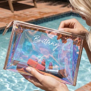 May include: A clear, iridescent cosmetic bag with a rose gold zipper and trim. The bag contains makeup items and the name "Brittany" is written on the front in white script. The bag is held near a swimming pool.
