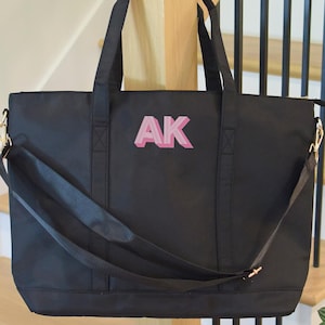 May include: Black tote bag with a pink monogram "AK" on the front. The bag has a long strap and two shorter handles.
