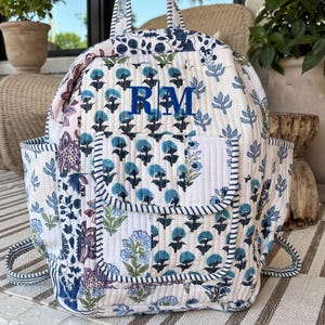 May include: A quilted backpack featuring a white base with a blue and green floral block print pattern. The bag includes a top handle, two side pockets, a front pocket with a flap, and the embroidered blue monogram letters RM on the front.