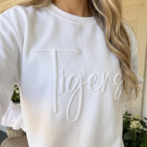 May include: A white long-sleeve crewneck sweatshirt featuring the word Tigers embossed in a raised, tonal white script font across the chest. The garment has a classic fit with ribbed cuffs and a ribbed hem, providing a clean, monochromatic look.