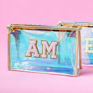 May include: Two iridescent clear plastic zippered pouches with gold trim. The pouch on the left has the letters "AM" in pink felt on the front. The pouch on the right has the letter "E" in white felt on the front.