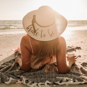 May include: A woman wearing a white sun hat with the text "bride to be" on it, sitting on a beach blanket with a drink in her hand.
