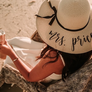 May include: A woman wearing a white dress and a straw sun hat with the text "mrs. price" on it. She is sitting on a blanket on the beach and holding a rose gold tumbler.