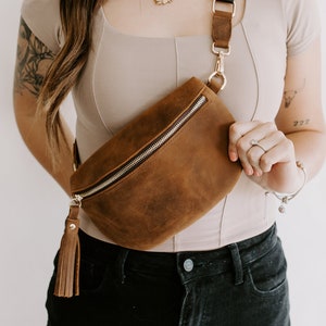 May include: A brown leather fanny pack with a gold zipper and a tassel. The bag is worn across the body with a black adjustable strap.