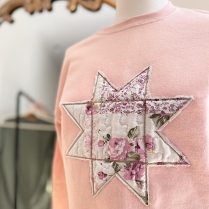 May include: A light pink crewneck sweatshirt featuring a large, eight-pointed star appliqué on the chest. The star is constructed from quilted fabric patches with a floral pattern in shades of pink, white, and green, finished with visible stitching.