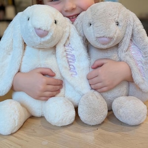 May include: Two plush bunny rabbits in cream and tan colors. The cream bunny has the name "Lillian" embroidered in purple on its ear. The bunnies have long floppy ears, pink noses, and are being held by a child.