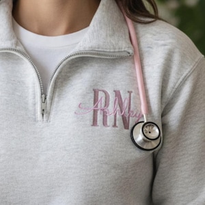 May include: A light gray quarter-zip sweatshirt with the embroidered letters "RN" in a cursive font. A pink stethoscope hangs around the neck. A white tumbler with a black lid is held in the hands.
