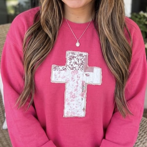 May include: A bright pink long-sleeve crewneck sweatshirt featuring a large, centered cross applique. The cross is constructed from various fabric patches in shades of white, light pink, and red, showcasing floral and checkered patterns.