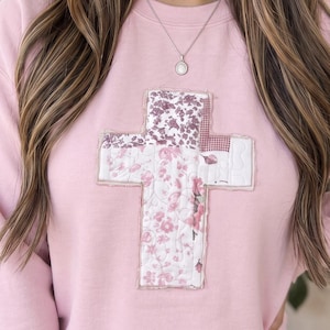 May include: A light pink crewneck sweatshirt featuring a large, centered cross applique. The cross is constructed from various quilted fabric patches in shades of pink, white, and mauve, showcasing floral and geometric patterns with visible stitching.