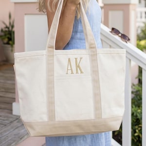 May include: A large off-white canvas tote bag featuring two sturdy shoulder straps and a reinforced base in a light tan color. The front of the bag displays the embroidered monogram letters AK in a matching tan thread centered on the upper panel.