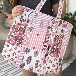 May include: A quilted tote bag featuring a patchwork design with pink and white floral patterns, vertical stripes, and small red hearts. The bag includes two fabric handles and a central panel with the embroidered letters KA in a matching pink thread.