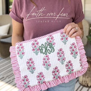 May include: A mauve t-shirt with the white text Faith over fear Isaiah 41:10 and a rectangular quilted pouch featuring a white background with a pink and green floral print, a green embroidered monogram, and pink and white striped ruffled edges.