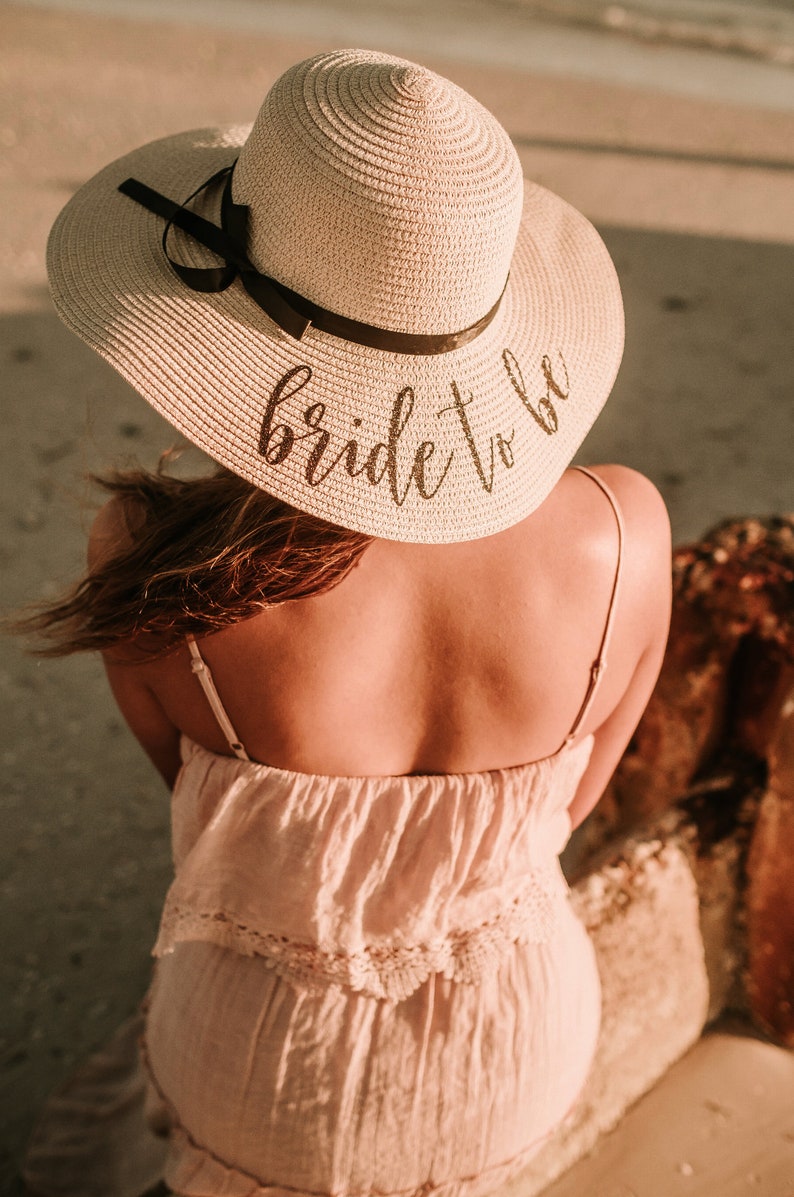 May include: A woman wearing a white straw sun hat with a black ribbon and the text "bride to be" in black script. She is wearing a white dress with lace trim.