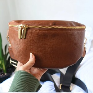 K&ouml;nnte beinhalten: Eine braune Leder-Bauchtasche mit einem goldenen Rei&szlig;verschluss und einem schwarzen verstellbaren Riemen.