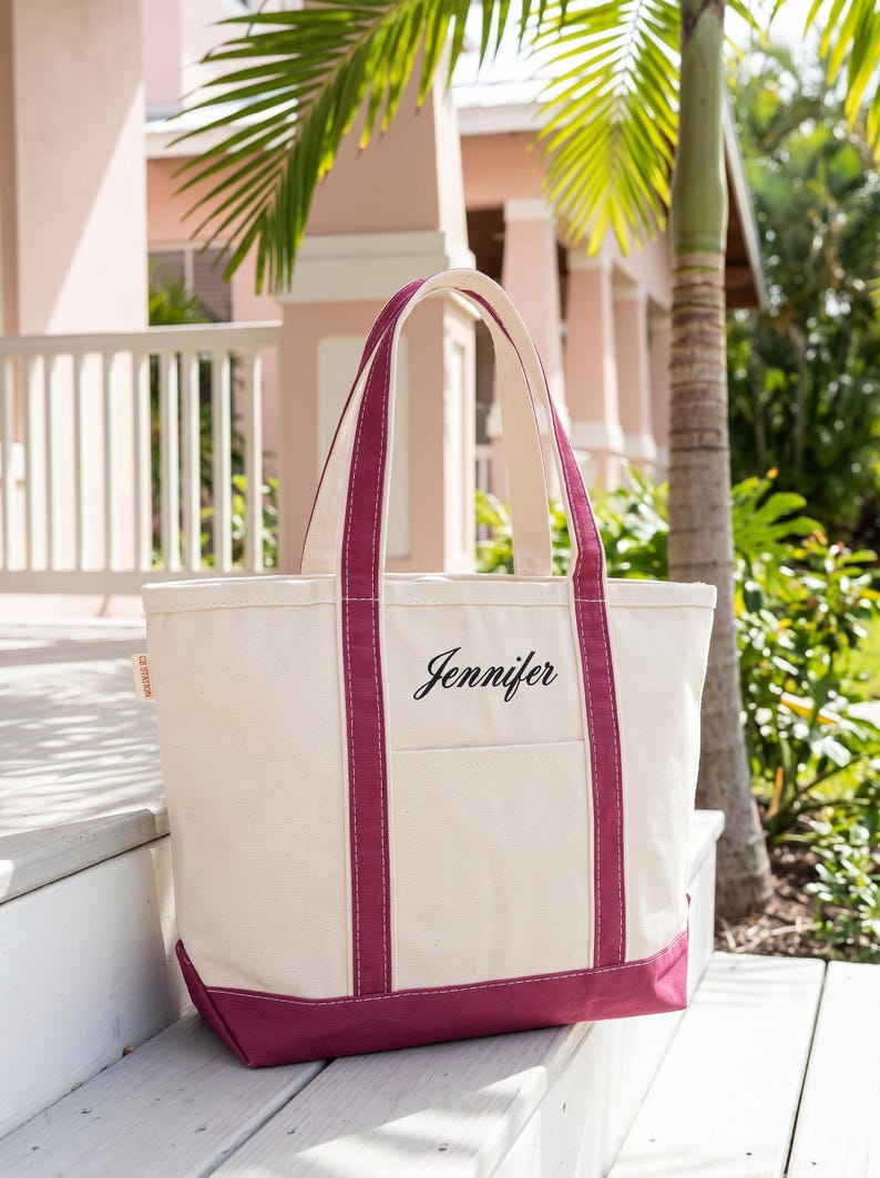 May include: Cream-colored canvas tote bag with a deep magenta base and accents. The bag features a front pocket and handles in magenta. The name "Jennifer" is elegantly scripted on the front. The bag is sitting on white steps.