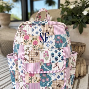 May include: A quilted backpack featuring a patchwork design of floral patterns, pink stripes, and brown gingham squares. The bag has a top handle, two side pockets, and a front flap pocket with the embroidered navy blue monogram letters SE.