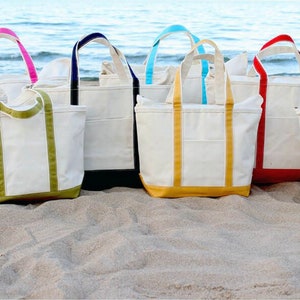 May include: A collection of canvas tote bags with various colored accents, including orange, pink, green, navy, yellow, red, and black. The bags are arranged on a sandy beach with the ocean in the background. Each bag has a matching colored bottom and handles.