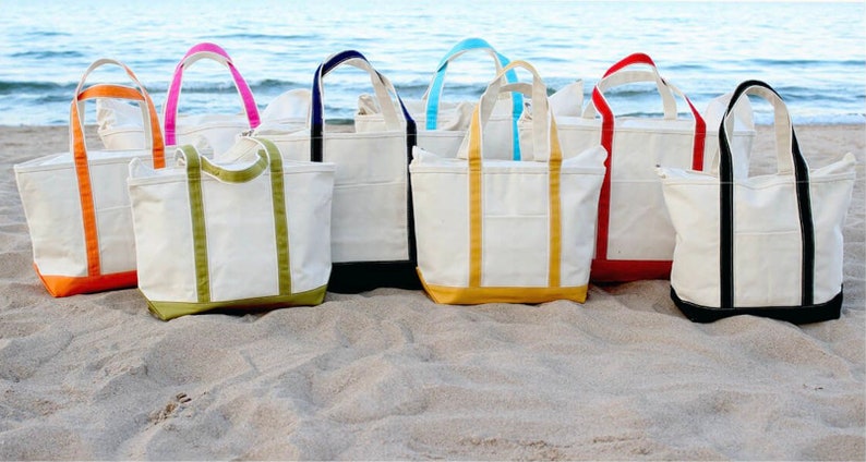 May include: A collection of canvas tote bags with various colored accents, including orange, pink, green, navy, yellow, red, and black. The bags are arranged on a sandy beach with the ocean in the background. Each bag has a matching colored bottom and handles.