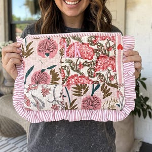 May include: A rectangular quilted pouch featuring a patchwork floral pattern in shades of pink, cream, and dark green. The pouch is bordered by a pink and white striped ruffle and includes the embroidered monogram LK and a small red heart.