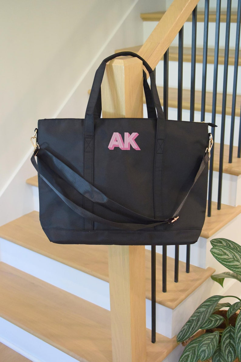 May include: Black tote bag with a pink monogram "AK" on the front. The bag has a long strap and two shorter handles.