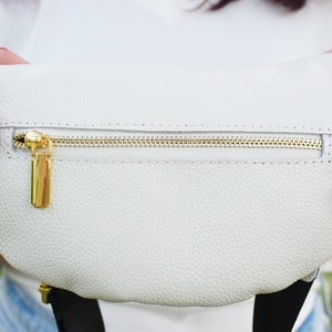 K&ouml;nnte beinhalten: Eine wei&szlig;e Leder-Bauchtasche mit einem goldenen Rei&szlig;verschluss und einem goldenen Rei&szlig;verschluss-Zipper. Die Bauchtasche wird von den H&auml;nden einer Person gehalten.