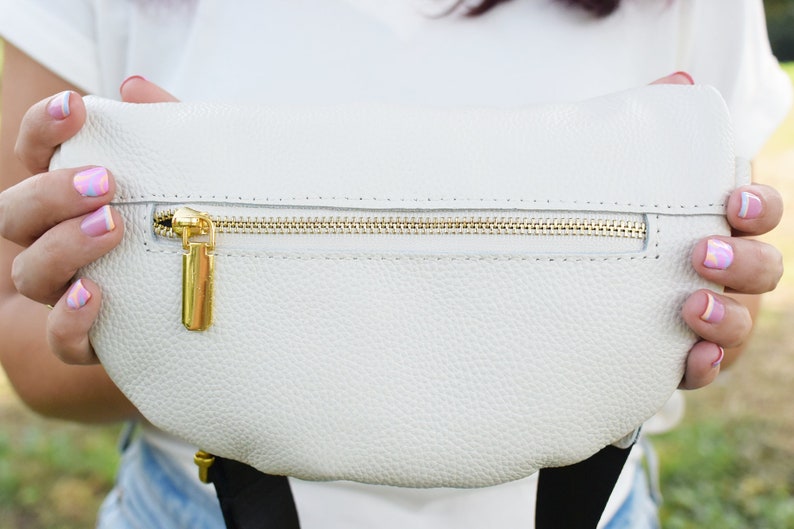 K&ouml;nnte beinhalten: Eine wei&szlig;e Leder-Bauchtasche mit einem goldenen Rei&szlig;verschluss und einem goldenen Rei&szlig;verschluss-Zipper. Die Bauchtasche wird von den H&auml;nden einer Person gehalten.