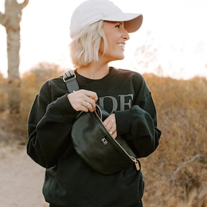 K&ouml;nnte beinhalten: Eine Frau tr&auml;gt ein schwarzes Sweatshirt mit dem Wort "IDE" darauf, eine wei&szlig;e Baseballkappe und eine schwarze Leder-Bauchtasche mit den Initialen "KB" darauf.