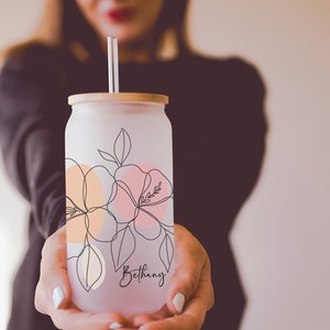 K&ouml;nnte beinhalten: Glasbecher mit Milchglas-Effekt, Bambusdeckel und klarem Strohhalm. Der Becher hat ein Blumendesign in Pfirsich, Rosa und Schwarz. Der Name "Bethany" steht in Schreibschrift auf der Vorderseite.