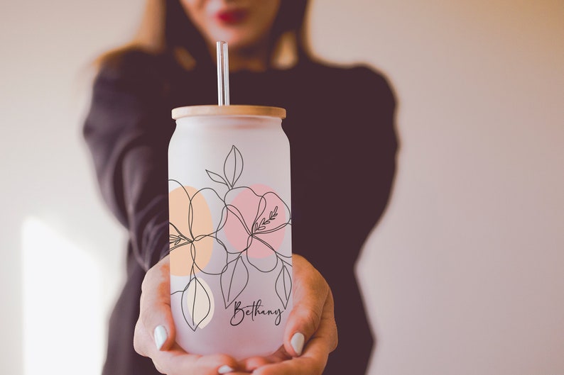 K&ouml;nnte beinhalten: Glasbecher mit Milchglas-Effekt, Bambusdeckel und klarem Strohhalm. Der Becher hat ein Blumendesign in Pfirsich, Rosa und Schwarz. Der Name "Bethany" steht in Schreibschrift auf der Vorderseite.