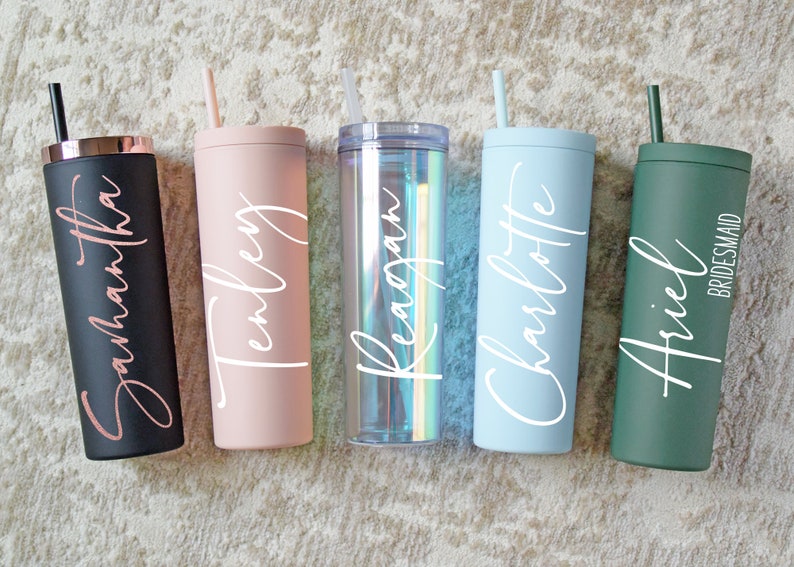 May include: Five personalized stainless steel tumblers with straws. The tumblers are in various colors, including black, pink, clear, blue, and green. Each tumbler has a name written on it in white lettering. The tumblers are arranged in a row on a beige surface.