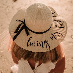 May include: A white straw sun hat with a black ribbon and the text "living my best" written in black script.