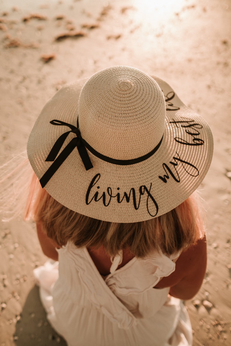 May include: A white straw sun hat with a black ribbon and the text "living my best" written in black script.