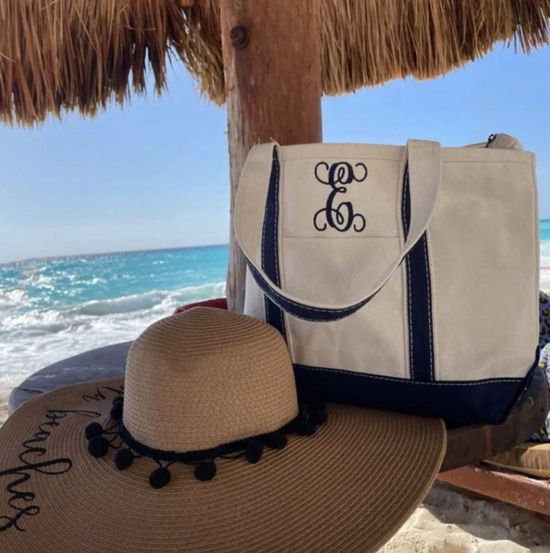 May include: A beige tote bag with navy blue trim and a monogram. A wide-brimmed straw hat with black pom-poms and the text "beaches" is in the foreground. The background shows the ocean and a wooden structure.