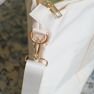 May include: A cream-colored tote bag with a gold zipper and a gold hook and loop strap. The bag has a cream-colored strap with a gold buckle.