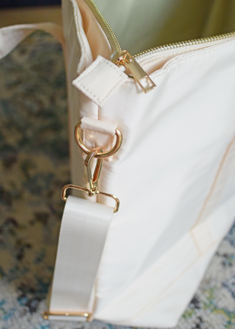May include: A cream-colored tote bag with a gold zipper and a gold hook and loop strap. The bag has a cream-colored strap with a gold buckle.