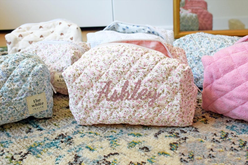 May include: A set of six quilted makeup bags with floral patterns. The bags are in various colors, including pink, blue, and white. One bag has the name "Ashley" embroidered on it in pink thread. The bags are all lined with a white fabric and have a zipper closure.