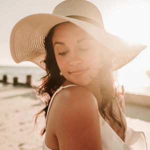 May include: A woman wearing a white sun hat and a white dress with ruffles. She is standing on a beach with the sun shining behind her.